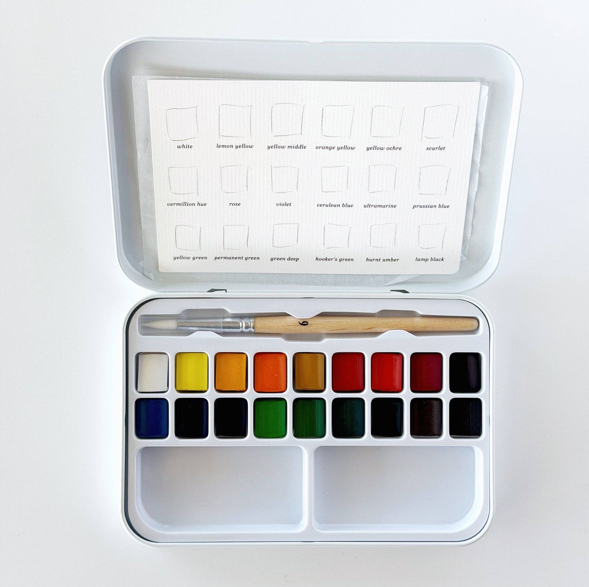 Emily Lex Studio watercolor paint set open flat on white surface; tin lid shows color swatch card with 18 labeled colors: white, lemon yellow, yellow middle, orange yellow, yellow ochre, scarlet, vermillion hue, rose, violet, cerulean blue, ultramarine, prussian blue, yellow green, permanent green, green deep, hooker's green, burnt umber, and lamp black; tin base holds 18 paint pans in two rows with two mixing trays below and a size 6 round brush resting across the top