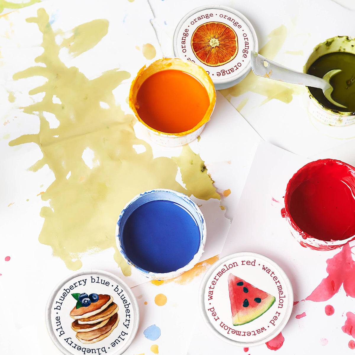Top-down flat lay of four open Elseware Unplug finger paint tubs with paint smears on white surface — orange, green, blue, and red liquid paints in tubs; illustrated food-themed lids beside each tub including orange slice, blueberry pancake, and watermelon wedge