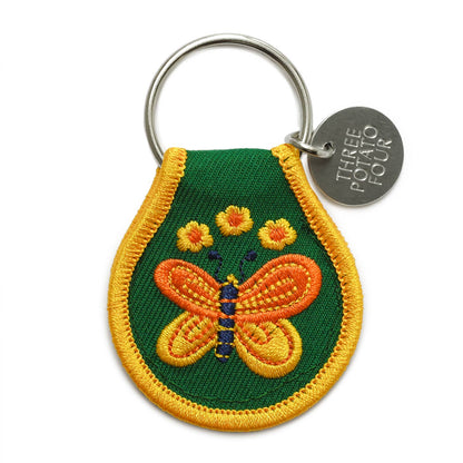 Three Potato Four Butterfly patch keychain on white background — teardrop-shaped natural cotton patch with forest green ground and gold merrowed border; embroidered orange and gold butterfly with navy-striped body in center; four small yellow daisy flowers above; silver metal split keyring at top with round debossed Three Potato Four brand charm hanging to the right