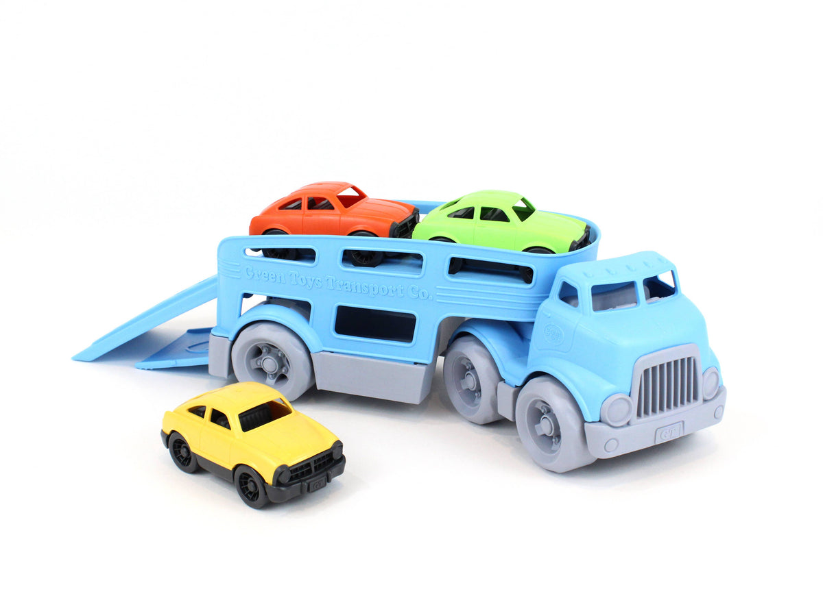 Green Toys Car Carrier on white background — blue double-decker transport truck viewed from the rear-left with the lower loading ramp extended diagonally; orange and green Mini Cars loaded on the upper deck; yellow Mini Car positioned in front of the ramp as if ready to drive up; six grey wheels and detachable trailer connection point visible
