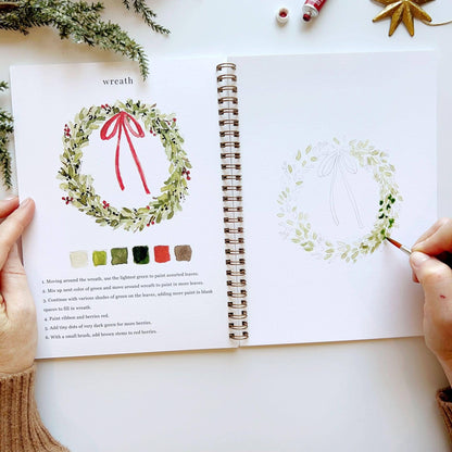 Open spread of Christmas Watercolor Workbook open to wreath page, hands holding the book showing completed green holly wreath with red bow and ribbon on left with color palette and step-by-step instructions, hand beginning to paint light green leaves on a wreath sketch on right, pine branches and gold star ornament in background