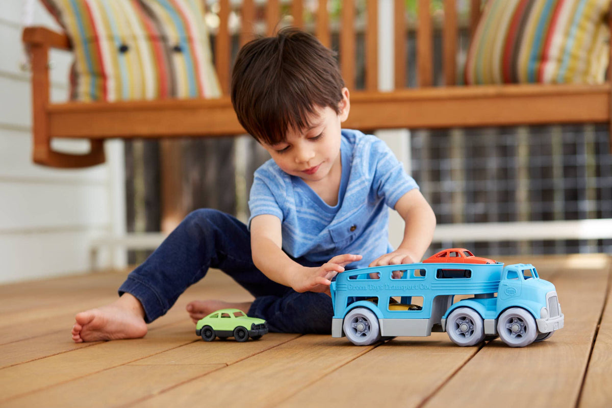 Green Toys Car Carrier on white background — blue double-decker transport truck viewed from the rear-right showing the back of the enclosed cab, the full length of the double-decker trailer with side rails and open lower deck, and six grey wheels; green and orange Mini Cars loaded on the upper deck; clean rear three-quarter angle showing trailer depth and proportions