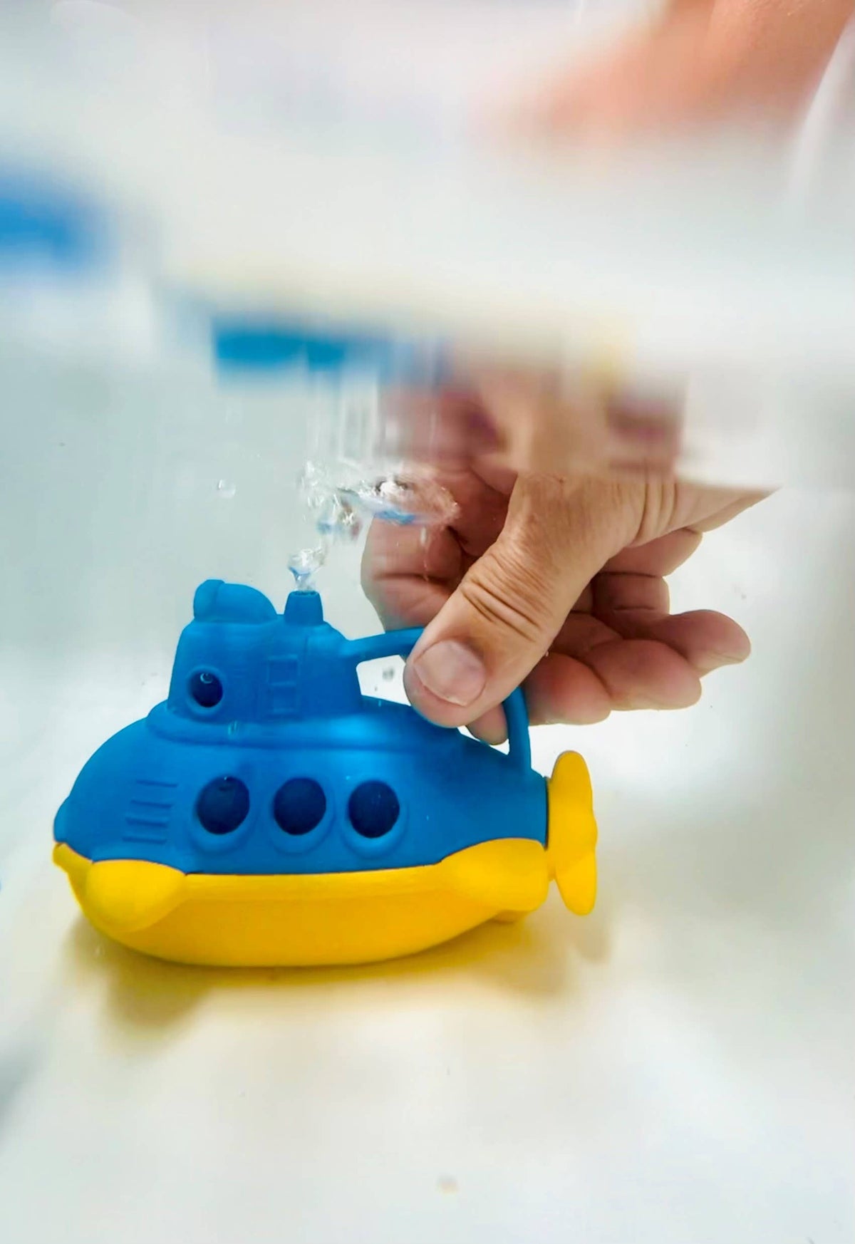 Close-up of an adult hand squeezing the handle of the Green Toys Bubbling Submarine in shallow water — blue and yellow submarine shown from the front-right with a stream of water shooting up from the blue periscope opening; water surface and light blue background visible above