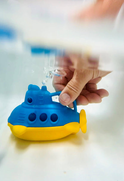 Close-up of an adult hand squeezing the handle of the Green Toys Bubbling Submarine in shallow water — blue and yellow submarine shown from the front-right with a stream of water shooting up from the blue periscope opening; water surface and light blue background visible above