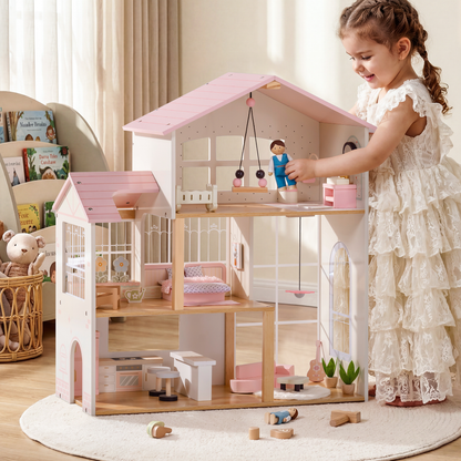 Young girl in white lace dress smiling while playing with a 3-story pink and cream wooden Robud dollhouse on a round rug in a warmly lit playroom — top floor children's room has a hanging mobile and wooden boy doll; middle floor has pink bedding and flower decor; ground floor shows kitchen appliances and stools; wooden doll figures and accessories scattered on the rug beside a bookshelf in the background
