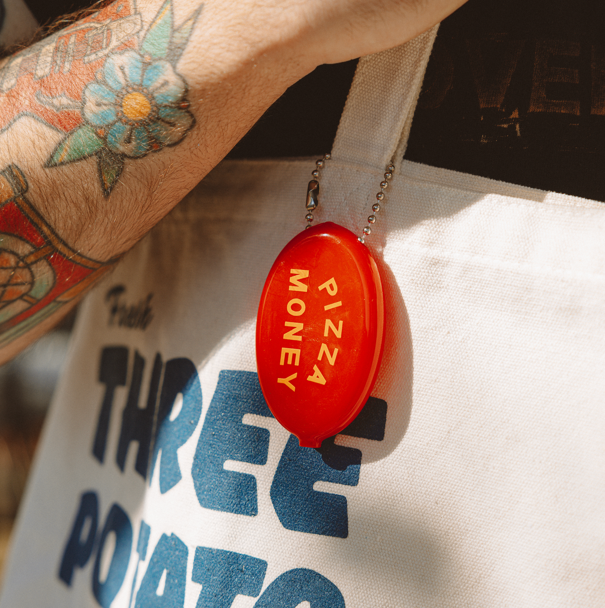 Three Potato Four Pizza Money coin pouch held by ball chain against a Three Potato Four canvas tote bag worn by a person with a floral tattoo on their wrist — red oval pouch hanging vertically showing "PIZZA MONEY" in orange lettering; blue block-letter Three Potato Four branding visible on the white tote in background