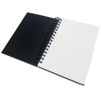 Elseware Unplug watercolor paper pad open at an angle on white background — black cover on the left; right side shows blank white cold press watercolor paper with prominent cotton rag texture; black spiral binding visible along the top edge