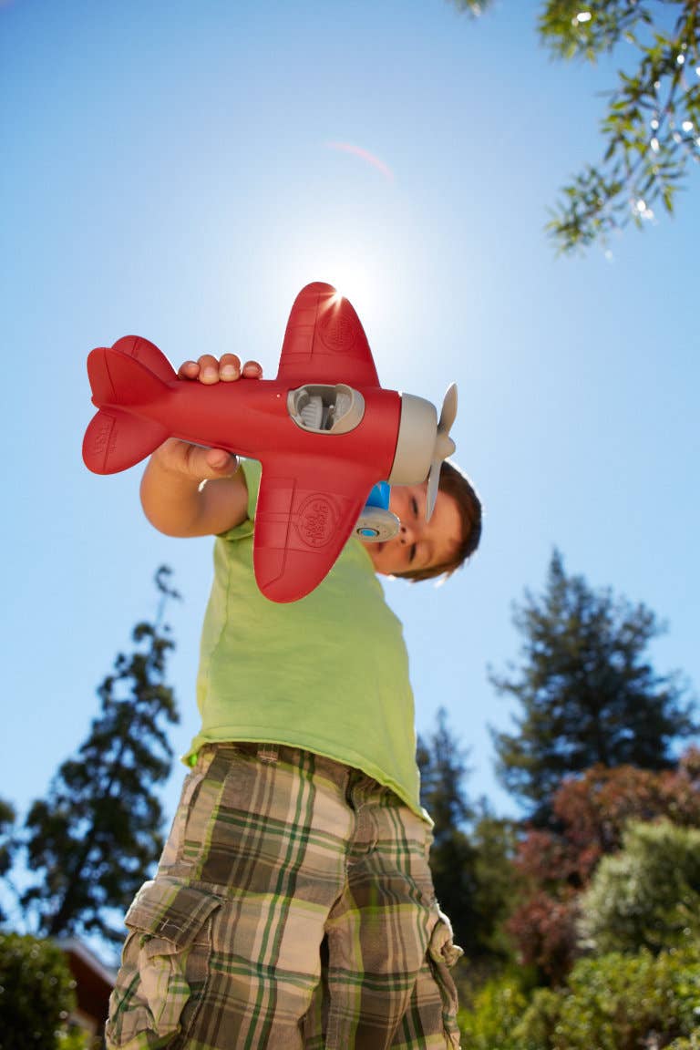 Young boy holding the Green Toys red airplane above his head outdoors on a bright sunny day — low-angle shot looking up at the child in a green t-shirt and plaid cargo shorts with blue sky, sun flare, and green trees in the background; the red plane with grey propeller and blue landing gear is held aloft with both hands as if in flight