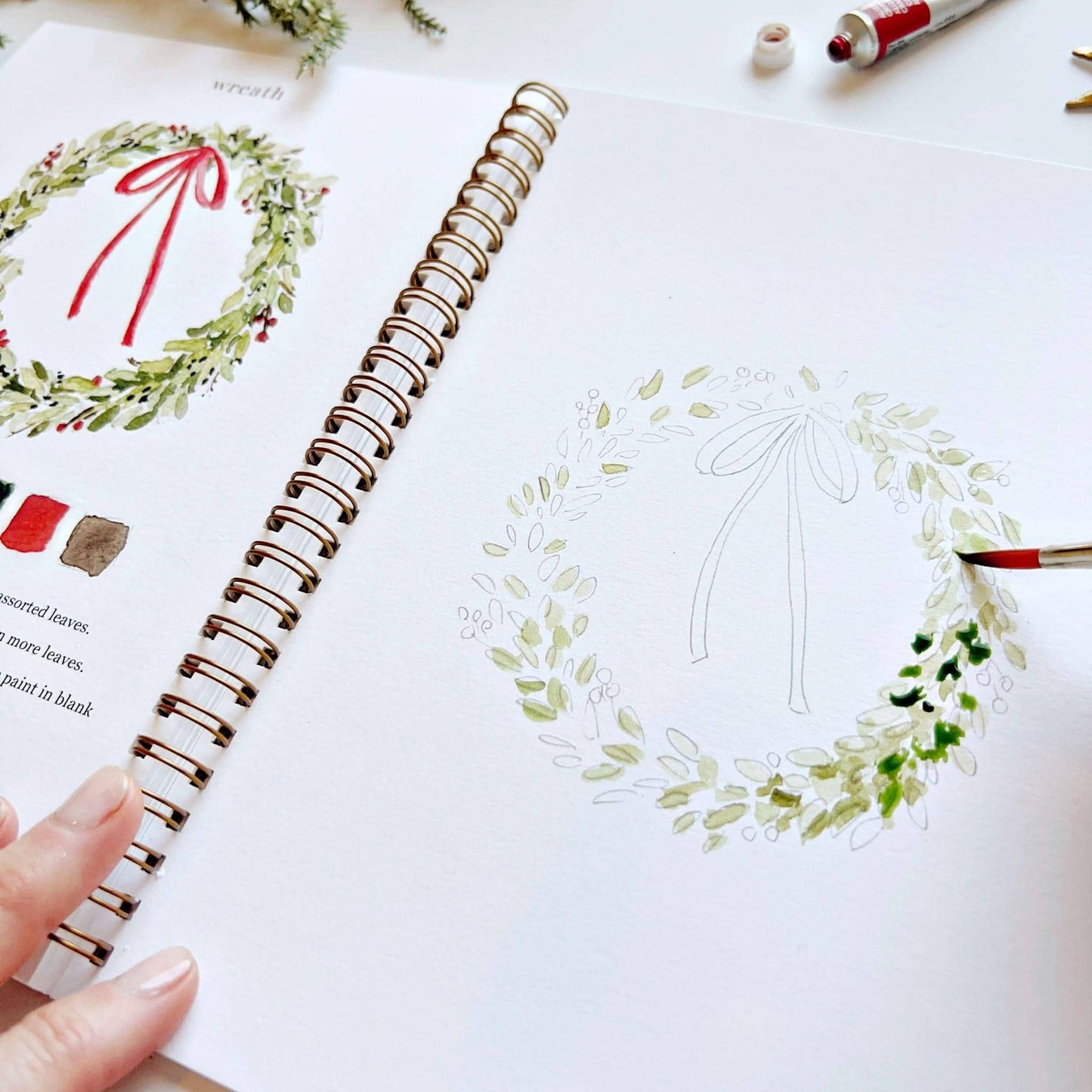 Angled close-up of Christmas Watercolor Workbook open to wreath page, hand adding darker green leaf and berry detail to a partially painted Christmas wreath sketch, with completed wreath illustration partially visible on left, watercolor tubes and gold star ornament in background