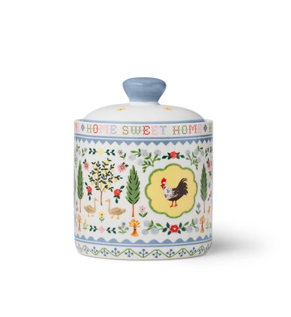 Rifle Paper Co. Home Sweet Home Porcelain Canister — front view — white porcelain canister with fitted dome lid and blue ceramic knob; wrap-around folk art pastoral illustration featuring a rooster in a yellow scalloped medallion at center, two geese, fruit trees with roses, tall cypress trees, wildflowers, and wheat sheaves; "HOME SWEET HOME" in red cross-stitch lettering around the neck; soft blue zigzag border bands; white background