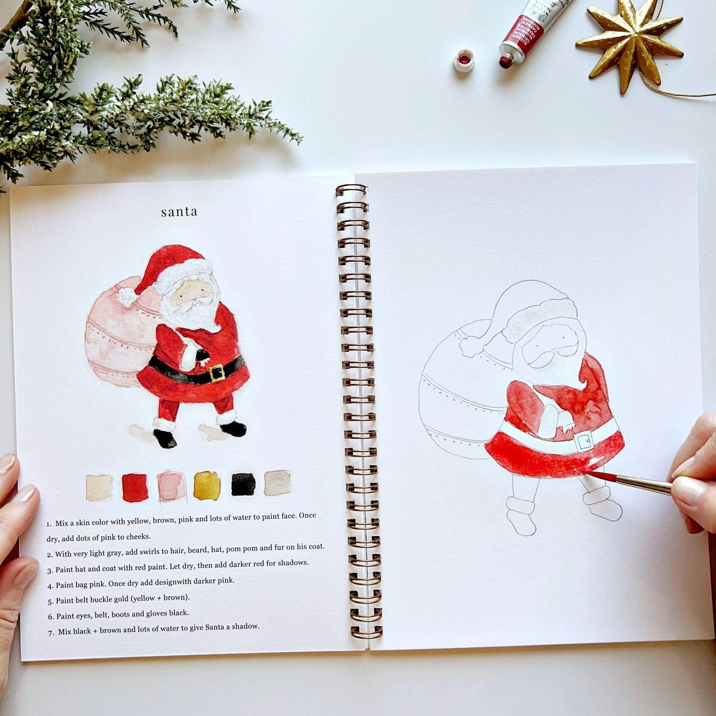Open spread of Christmas Watercolor Workbook open to Santa page, hands holding the book showing completed Santa with gift bag on left with color palette and step-by-step instructions, hand painting red watercolor onto a partially painted Santa sketch on right, pine branches and gold star ornament in background