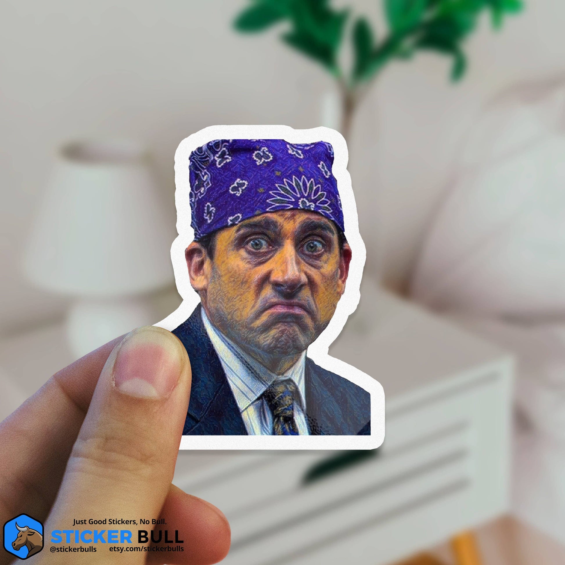 Hand holding a Sticker Bull Prison Mike The Office die cut vinyl sticker against a blurred living room background — illustrated Michael Scott as Prison Mike in purple bandana; white die cut border; Sticker Bull logo in corner