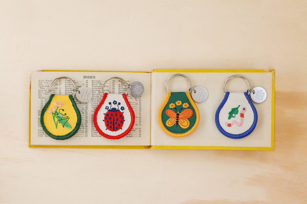 Four Three Potato Four insect patch keychains laid on open vintage book index pages inside a yellow-bordered book on cream surface — left to right: green Grasshopper keychain on yellow patch, red Ladybug keychain on white patch, orange Butterfly keychain on green patch, green Worm keychain on white patch with navy border; all shown with silver rings and debossed brand charms