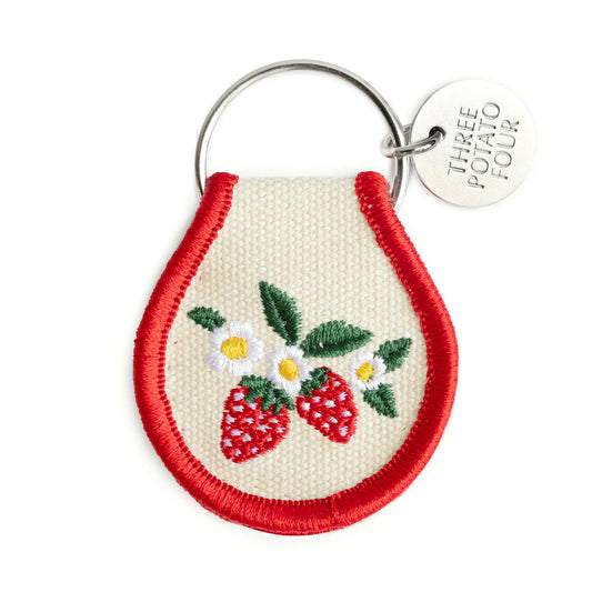 Three Potato Four Strawberry Blossom patch keychain on white background — rounded teardrop-shaped natural cream cotton patch with red merrowed border; embroidered design of two red strawberries with white seed dots flanked by white daisy blossoms with yellow centers and dark green serrated leaves; silver metal split keyring at top with round debossed Three Potato Four brand charm hanging to the right