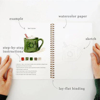 Flat lay of Christmas Watercolor Workbook open to an interior page with Christmas illustrations and step-by-step instructions