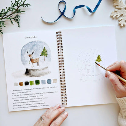 Open spread of Winter Watercolor Workbook open to snow globe page, hand beginning to paint green watercolor on the Christmas tree inside a snow globe sketch with a reindeer, with completed snow globe illustration, color palette, and step-by-step instructions on left, blue ribbon and snowflake ornament in background