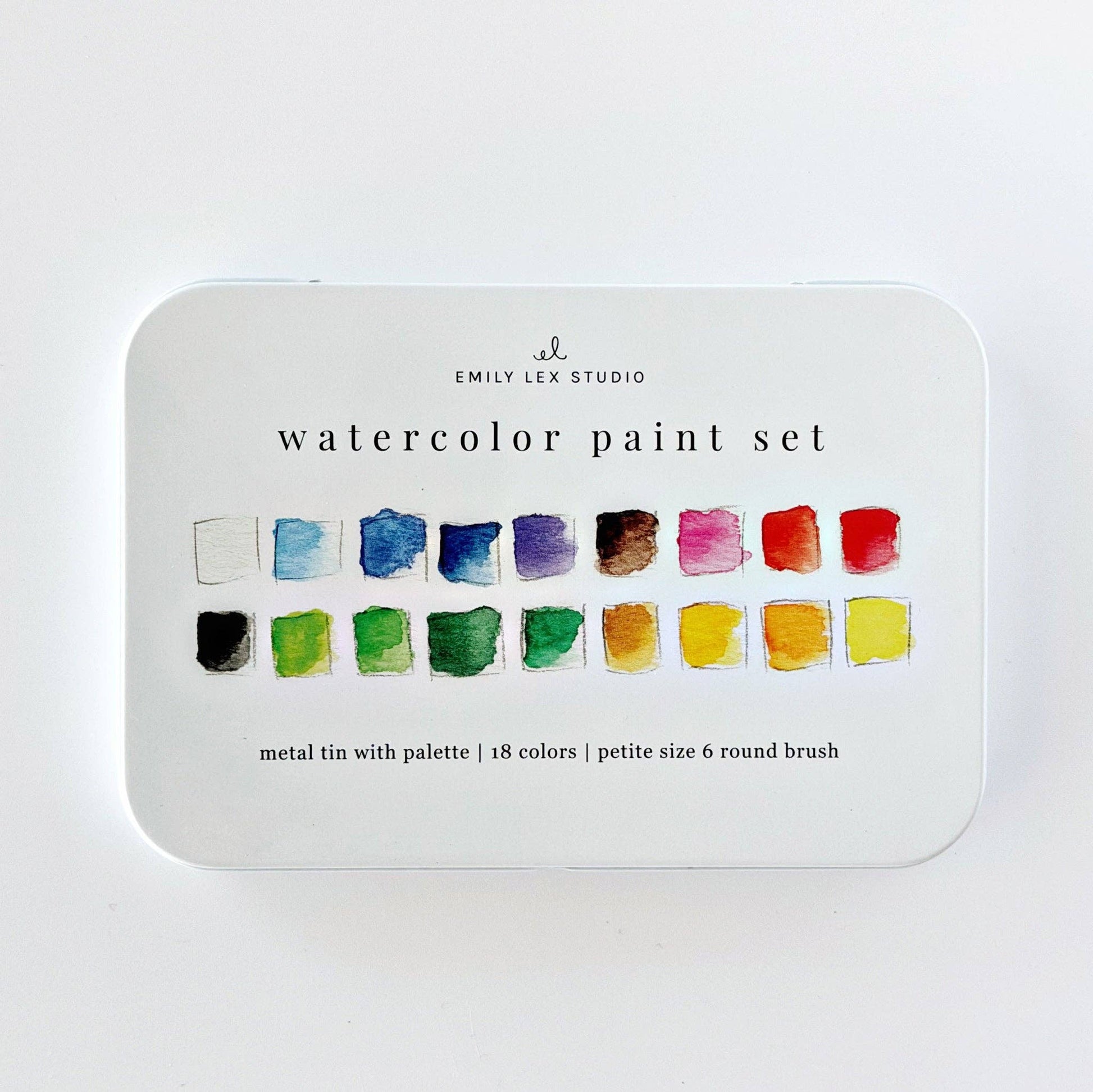 Emily Lex Studio watercolor paint set flat lay on white surface; open tin at upper right showing 18 paint pans in three rows with two mixing trays at bottom; color swatch card at lower left with all 18 colors labeled; size 6 round brush with wood handle resting diagonally; product box at upper left