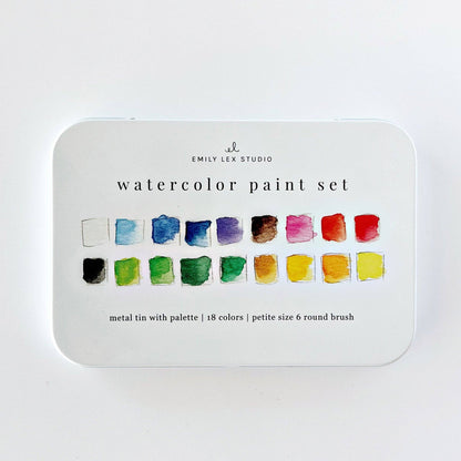 Emily Lex Studio watercolor paint set flat lay on white surface; open tin at upper right showing 18 paint pans in three rows with two mixing trays at bottom; color swatch card at lower left with all 18 colors labeled; size 6 round brush with wood handle resting diagonally; product box at upper left