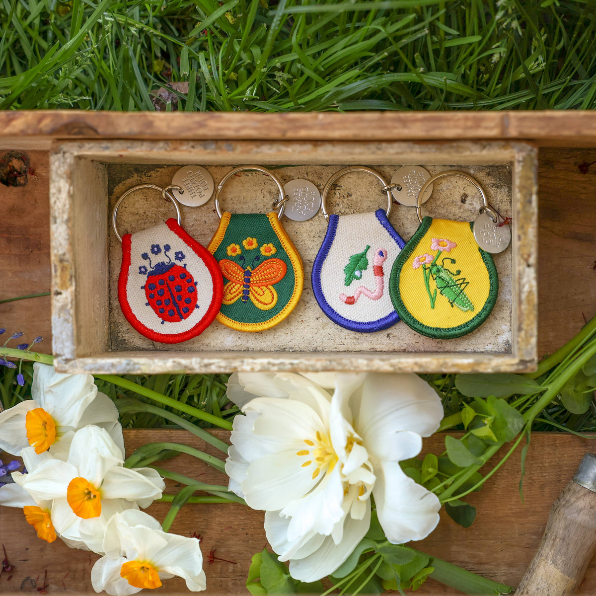Four Three Potato Four insect patch keychains displayed in a rustic wooden tray on a garden surface with grass and white daffodils — left to right: red-bordered Ladybug keychain on white patch, green-bordered Butterfly keychain on forest green patch, purple-bordered Worm keychain on white patch, yellow-bordered Grasshopper keychain on yellow patch; all with silver split rings and brand charms