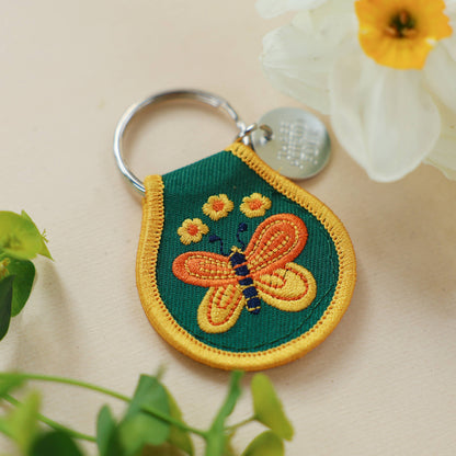 Three Potato Four Butterfly patch keychain on cream surface with flowers — teardrop patch shown at slight angle with green ground and gold border; embroidered orange butterfly with navy body and yellow daisy accents; silver split ring and Three Potato Four charm visible; white daffodils and green leaves in background