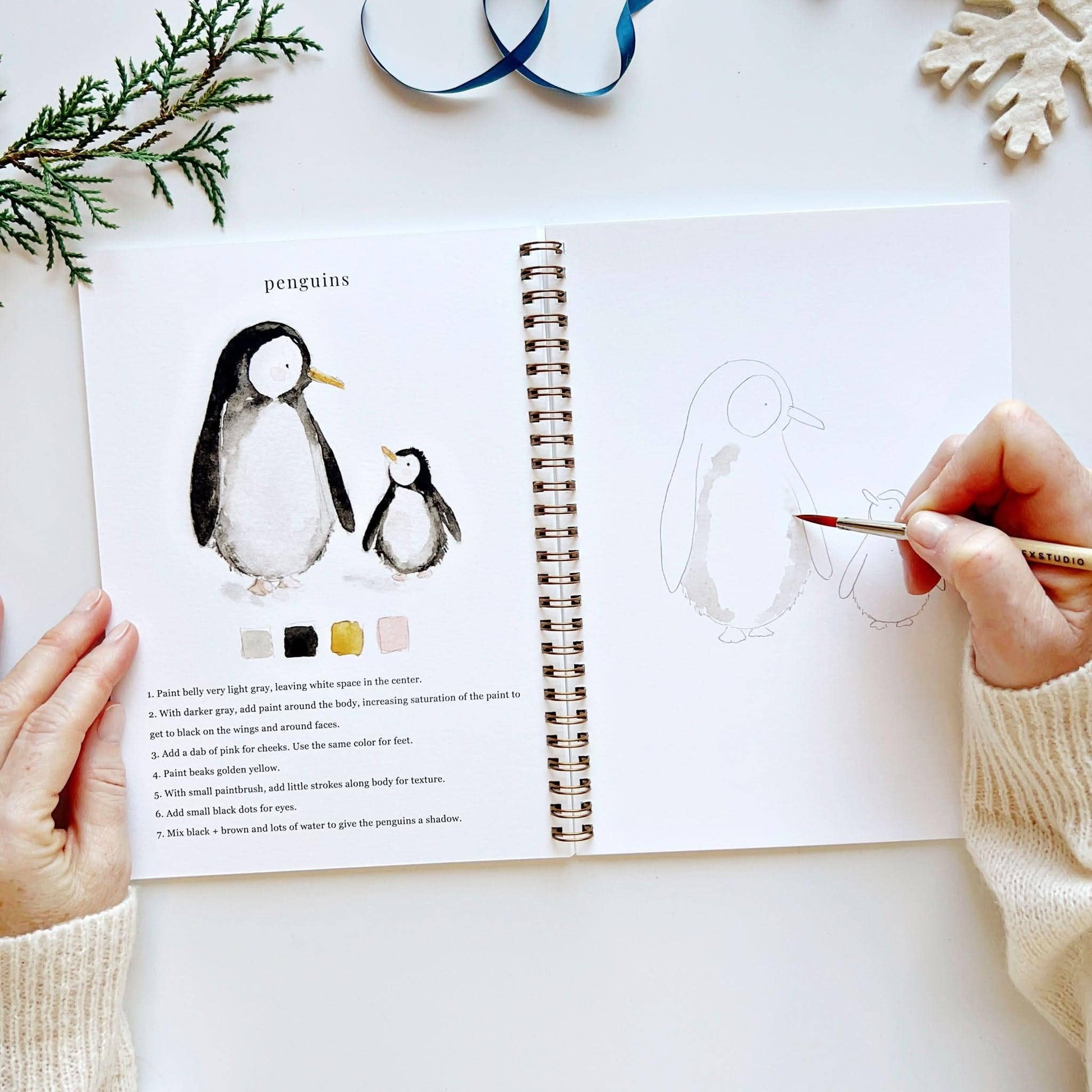 Open spread of Winter Watercolor Workbook open to penguins page, hand beginning to paint light gray watercolor on a large penguin sketch, with completed adult and baby penguin illustration, color palette, and step-by-step instructions on left, cedar branch, blue ribbon, and snowflake ornament in background