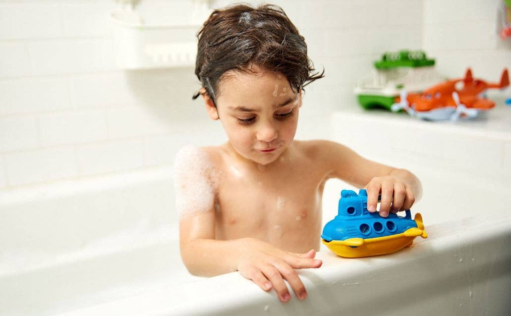 Young child with wet hair playing with the Green Toys blue and yellow Bubbling Submarine in a bubble bath — child leans over the edge of a white bathtub holding the submarine on the tub ledge with one hand; a green Green Toys boat and orange Green Toys airplane are visible blurred in the background on the tub ledge