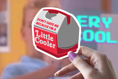 Hand holding a Sticker Bull Don't Hate Me Because I'm A Little Cooler die cut vinyl sticker in front of a blurred neon sign background reading "Very Cool" — illustrated red and gray mini cooler with red and white pun text; white die cut border; purple manicured nails

