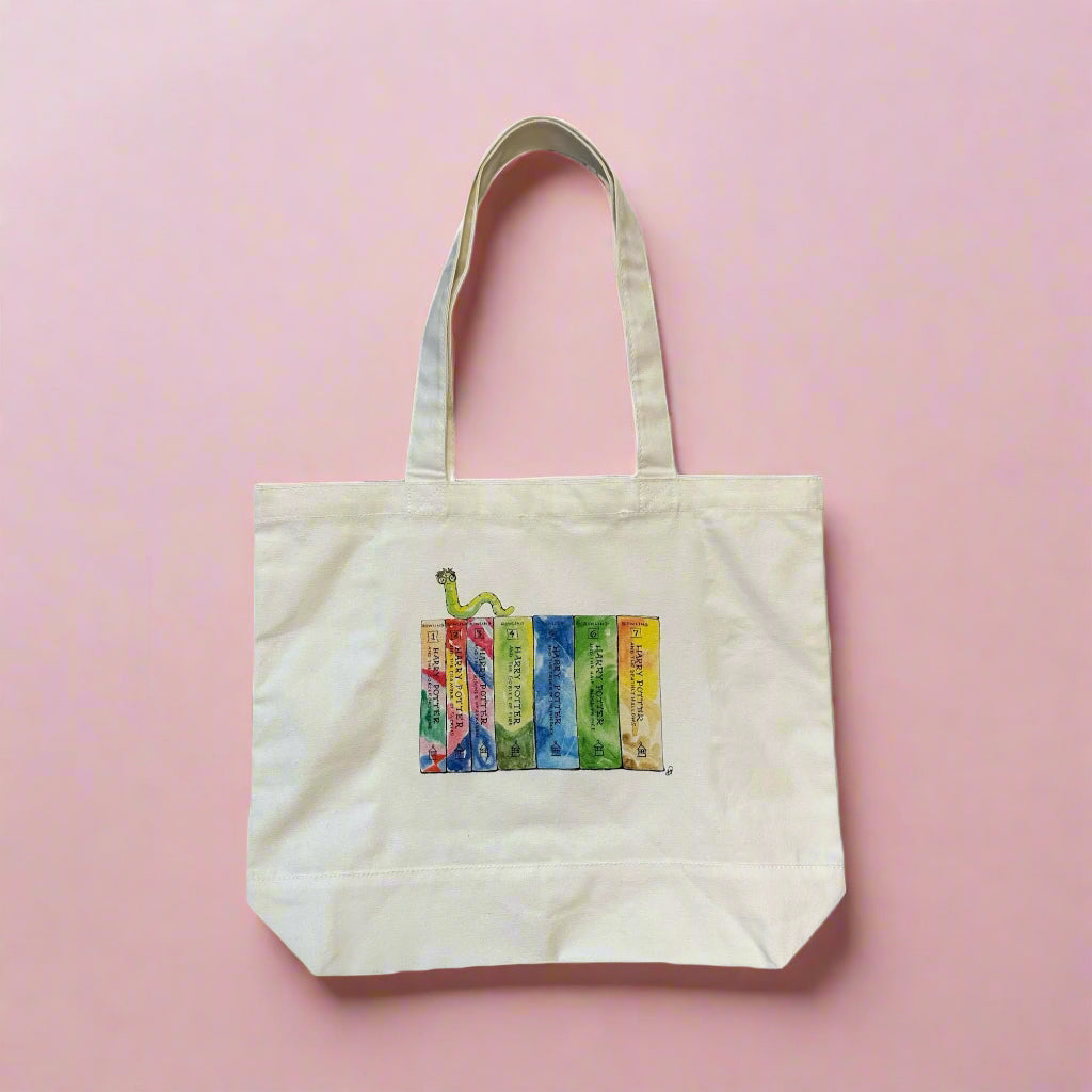 Natural canvas tote bag with watercolor Harry Potter book spine illustration on a pink background, full bag view showing handles and gusset