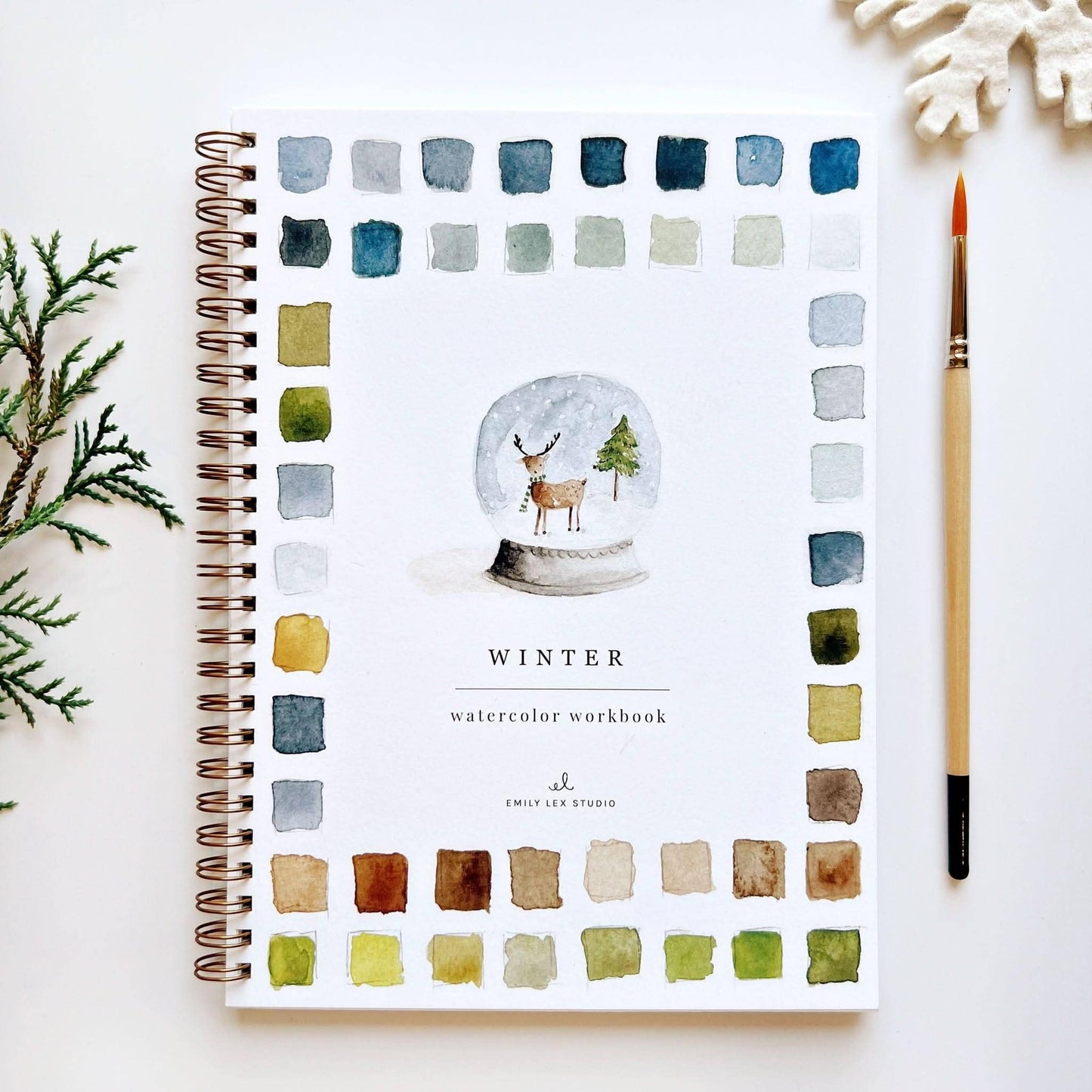 Winter Watercolor Workbook by Emily Lex Studio — spiral-bound beginner watercolor painting book with a snow globe illustration featuring a reindeer and Christmas tree on the cover, bordered by a grid of blue, green, and earth-tone watercolor swatches