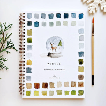 Winter Watercolor Workbook by Emily Lex Studio — spiral-bound beginner watercolor painting book with a snow globe illustration featuring a reindeer and Christmas tree on the cover, bordered by a grid of blue, green, and earth-tone watercolor swatches