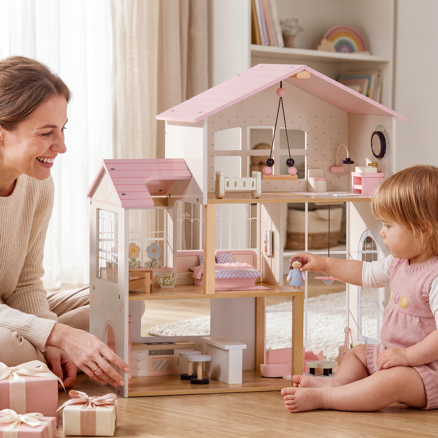 Smiling mother and toddler girl playing together with a Robud 3-story pink and cream wooden dollhouse on a wood floor — mother kneeling beside it with pink gift boxes nearby; toddler holding a wooden doll figure toward the middle floor; greenhouse tower visible on the left; bookshelf with rainbow art in background