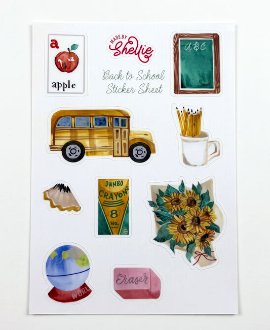 Made by Shellie Back to School Sticker Sheet — 9 watercolor die-cut stickers including "a is for apple" flashcard, green ABC chalkboard, yellow school bus, mug of pencils, paper airplane, jumbo crayons box, sunflower bouquet, world globe on red stand, and pink eraser; white sheet on white background