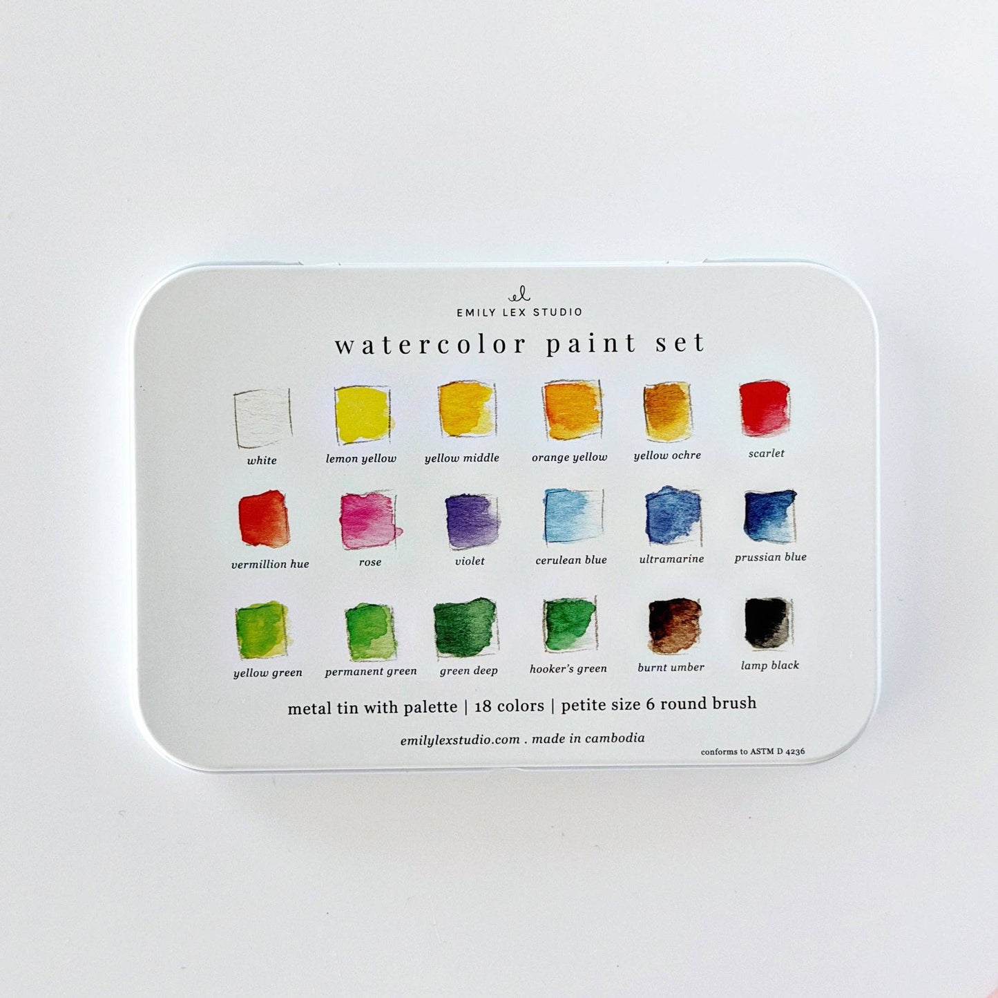 Emily Lex Studio watercolor paint set tin lid flat on white surface; lid shows all 18 colors labeled in three rows of 6: white, lemon yellow, yellow middle, orange yellow, yellow ochre, scarlet, vermillion hue, rose, violet, cerulean blue, ultramarine, prussian blue, yellow green, permanent green, green deep, hooker's green, burnt umber, lamp black; "metal tin with palette | 18 colors | petite size 6 round brush · emilylexstudio.com · made in cambodia · conforms to ASTM D 4236" text at bottom
