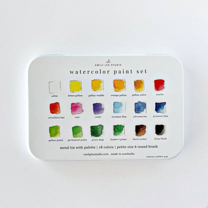 Emily Lex Studio watercolor paint set tin lid flat on white surface; lid shows all 18 colors labeled in three rows of 6: white, lemon yellow, yellow middle, orange yellow, yellow ochre, scarlet, vermillion hue, rose, violet, cerulean blue, ultramarine, prussian blue, yellow green, permanent green, green deep, hooker's green, burnt umber, lamp black; "metal tin with palette | 18 colors | petite size 6 round brush · emilylexstudio.com · made in cambodia · conforms to ASTM D 4236" text at bottom