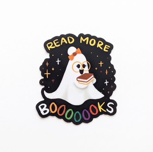 Read More Booooooks Ghost vinyl sticker by Little Tear Drop — die-cut sticker with black background; adorable white ghost wearing round gold glasses and an orange bow holds a small book; gold stars and cross sparkles surround the ghost; "READ MORE" arched in yellow text at top; "BOOOOOOKS" in multicolor letters at bottom; 2.75"×3"