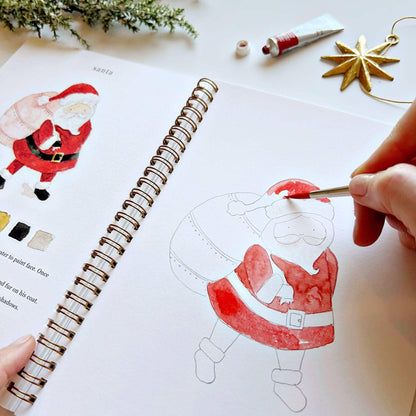 Angled close-up of Christmas Watercolor Workbook open to Santa page, hand painting red watercolor onto the coat of a partially painted Santa sketch, with completed Santa illustration and color palette partially visible on left, gold star ornament in background