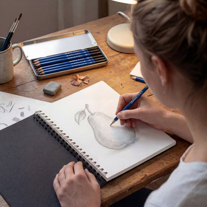 Lifestyle photo of adult woman sketching a pear in a spiral-bound sketchbook on a wooden desk — holding a blue Elseware Unplug 2B drawing pencil; open silver pencil tin with remaining 5 pencils visible in background beside a white mug of paintbrushes, eraser, pencil shavings, and warm lamp light