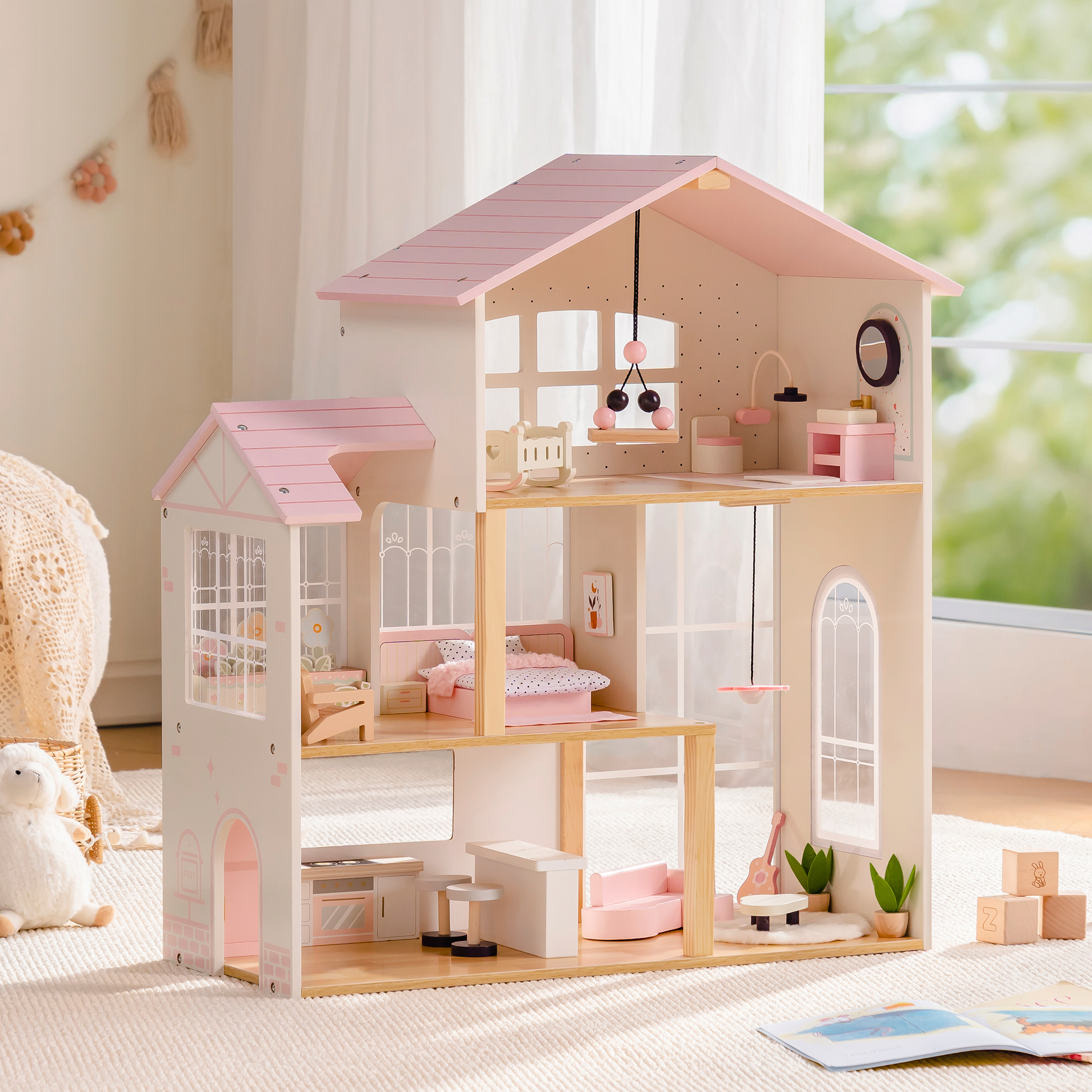 Robud 3-story pink and cream wooden dollhouse in a bright playroom — pink roof and open-front design showing top-floor nursery with hanging mobile, middle-floor bedroom with pink bedding, and ground-floor kitchen and living room with pink sofa; wooden doll figures and building blocks on the floor beside a woven basket
