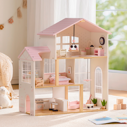 Robud 3-story pink and cream wooden dollhouse in a bright playroom — pink roof and open-front design showing top-floor nursery with hanging mobile, middle-floor bedroom with pink bedding, and ground-floor kitchen and living room with pink sofa; wooden doll figures and building blocks on the floor beside a woven basket
