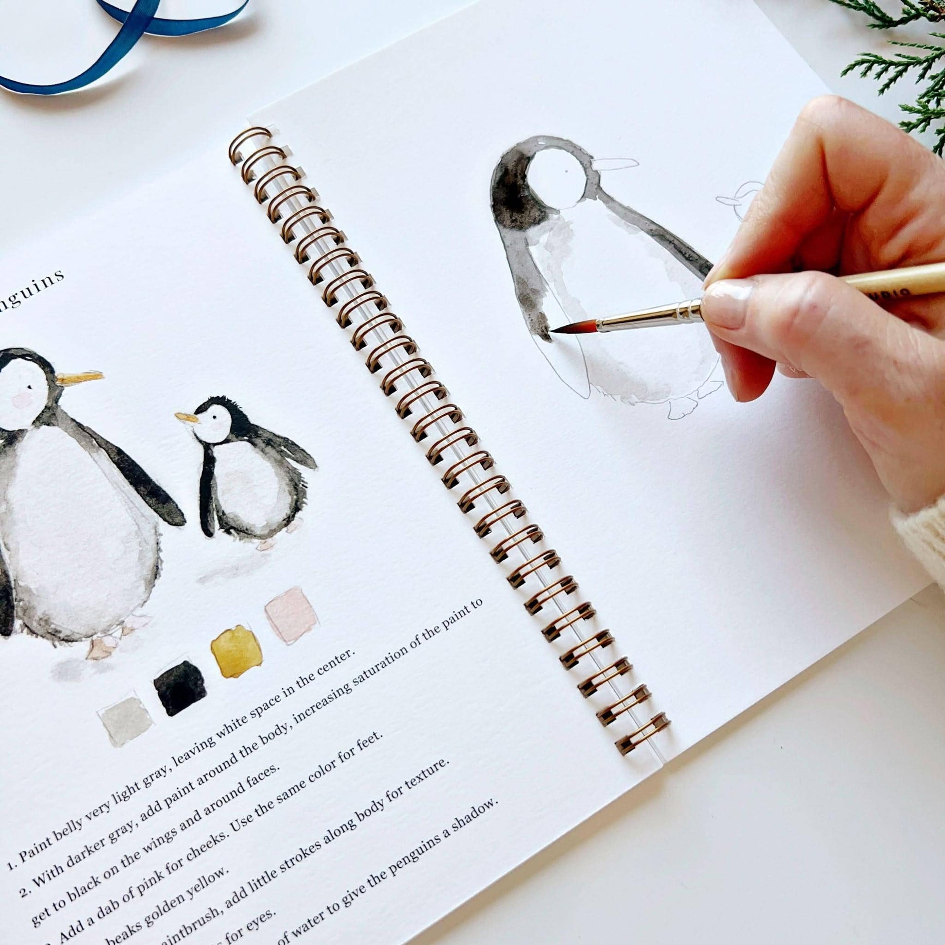 Angled close-up of Winter Watercolor Workbook open to penguins page, hand painting dark gray and black watercolor onto the wings of a partially painted penguin sketch, with completed penguin illustrations and color palette partially visible on left, blue ribbon and cedar branch in background