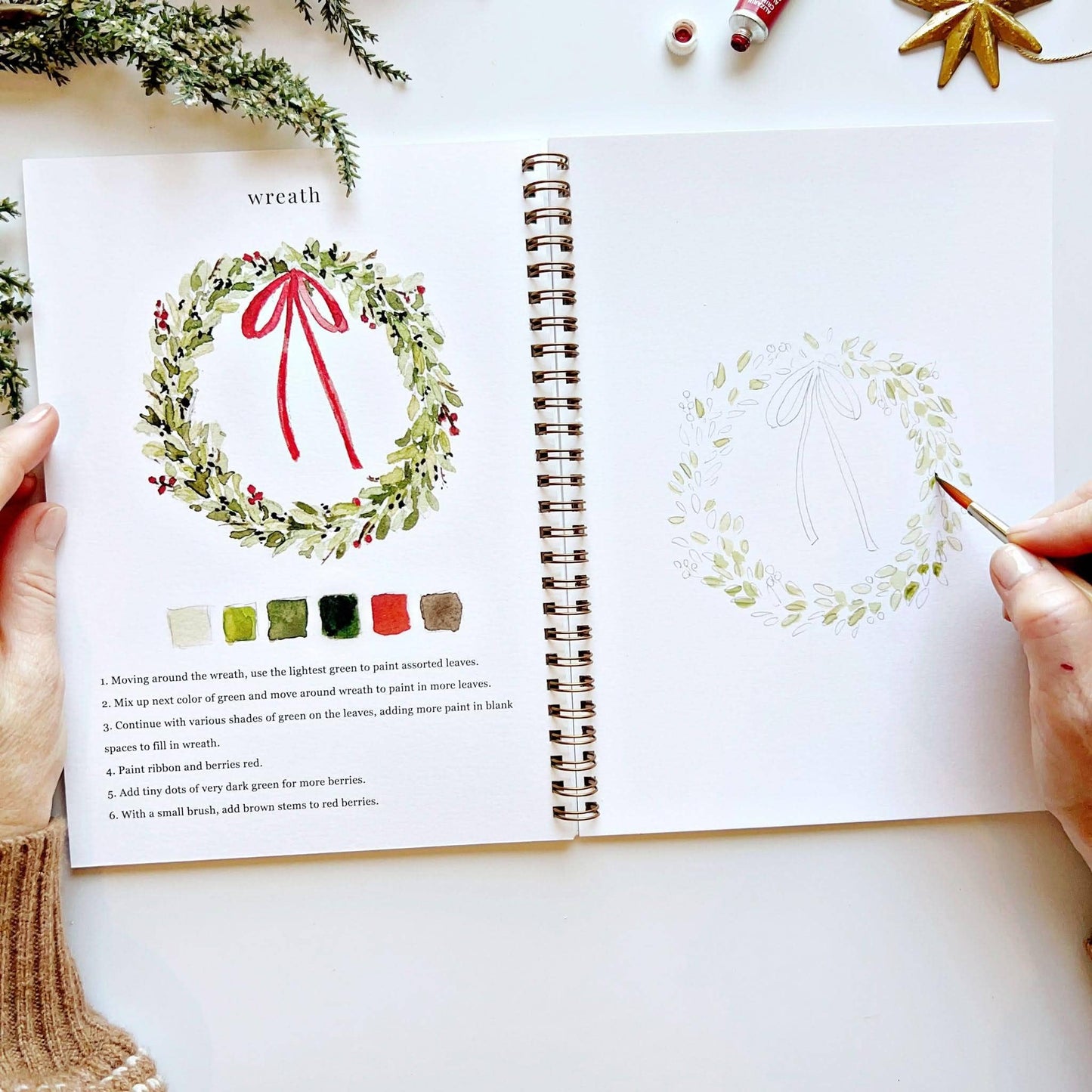 Overhead view of Christmas Watercolor Workbook open to wreath page, hand painting green leaves on a partially painted Christmas wreath sketch with red bow, with completed wreath illustration and color palette on left, gold star ornament in background