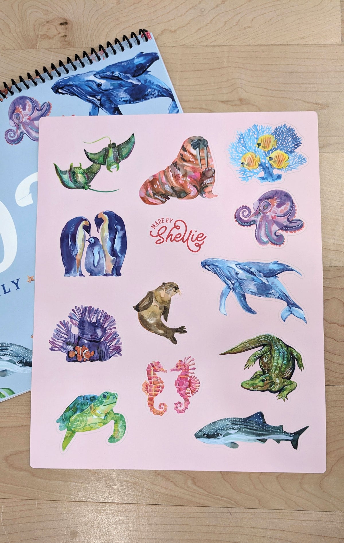 Made by Shellie Sea Life Sticker Sheet flat on a wood surface next to the Sea Life spiral calendar, close-up showing all 12 watercolor sea creature stickers in full detail on blush pink sheet