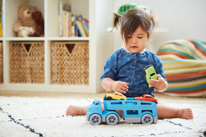 Toddler girl with pigtails in a blue denim top sitting cross-legged on a white shag rug playing with the Green Toys Car Carrier — she holds a green Mini Car in her right hand and reaches toward the blue double-decker carrier truck on the floor in front of her; yellow and red Mini Cars loaded on the carrier; white bookshelf with stuffed bear and woven baskets and a striped floor cushion visible in the background