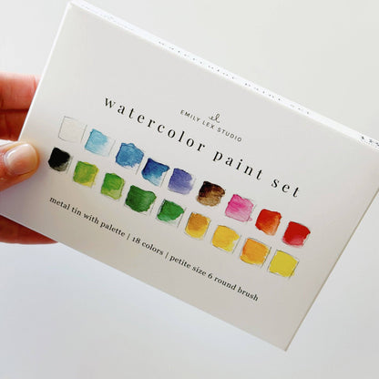Hand holding the Emily Lex Studio watercolor paint set box; front of box shows 18 watercolor swatches in two rows and "watercolor paint set · metal tin with palette | 18 colors | petite size 6 round brush" text; Emily Lex Studio logo at top