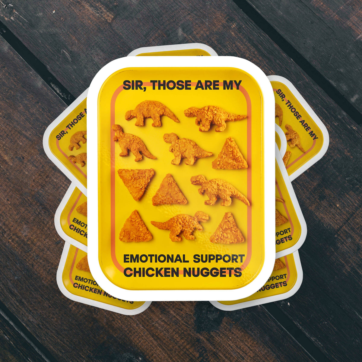 Stack of Sticker Bull Sir, Those Are My Emotional Support Chicken Nuggets die cut vinyl stickers fanned out on a dark wood surface — yellow tray with dino nuggets design and bold black text; white die cut border