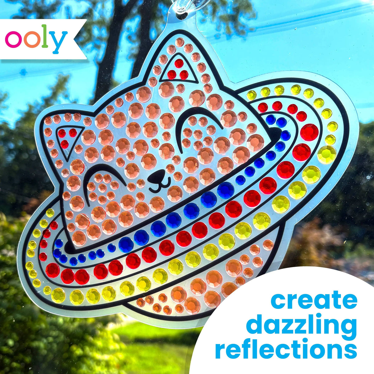 Completed OOLY Sun Sparkles space cat with planet ring gem suncatcher hanging in a sunny window, decorated with orange, red, blue, and yellow gems — OOLY logo and text "create dazzling reflections"