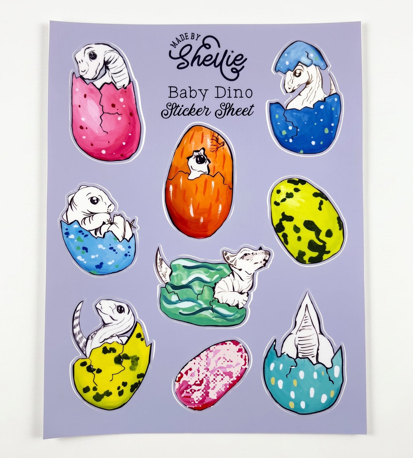 Made by Shellie Baby Dino Sticker Sheet — 9 die-cut stickers of white line-art baby dinosaurs hatching from colorful speckled eggs in pink, blue, orange, yellow-green, and teal; periwinkle blue sheet; white background