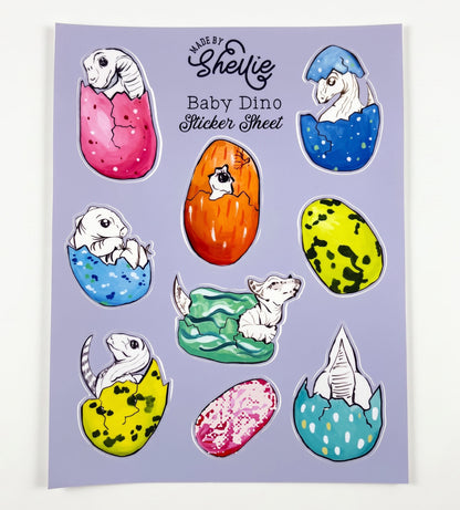 Made by Shellie Baby Dino Sticker Sheet — 9 die-cut stickers of white line-art baby dinosaurs hatching from colorful speckled eggs in pink, blue, orange, yellow-green, and teal; periwinkle blue sheet; white background