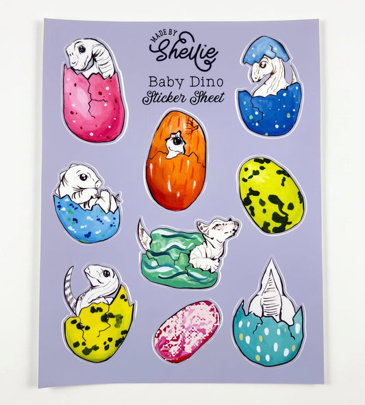 Made by Shellie Baby Dino Sticker Sheet — 9 die-cut stickers of white line-art baby dinosaurs hatching from colorful speckled eggs in pink, blue, orange, yellow-green, and teal; periwinkle blue sheet; white background