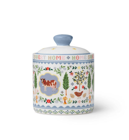 Rifle Paper Co. Home Sweet Home Porcelain Canister — back view — reverse side showing a brown and white cow in a blue scalloped medallion at center, a hen with eggs, fruit trees, cypress trees, roses, and wildflowers; "SWEET HOME" cross-stitch lettering visible around neck; soft blue zigzag border bands; white background